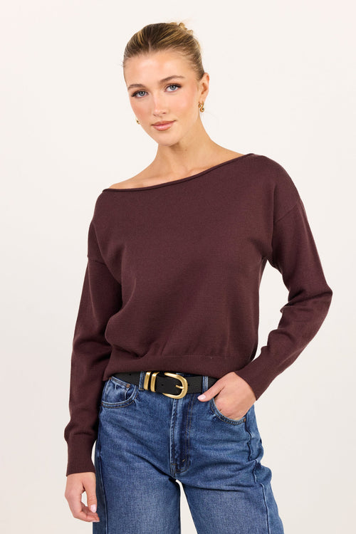 A woman modeling a dark brown off-the-shoulder knit sweater with blue jeans and a black belt with a gold buckle, set against a plain light background.