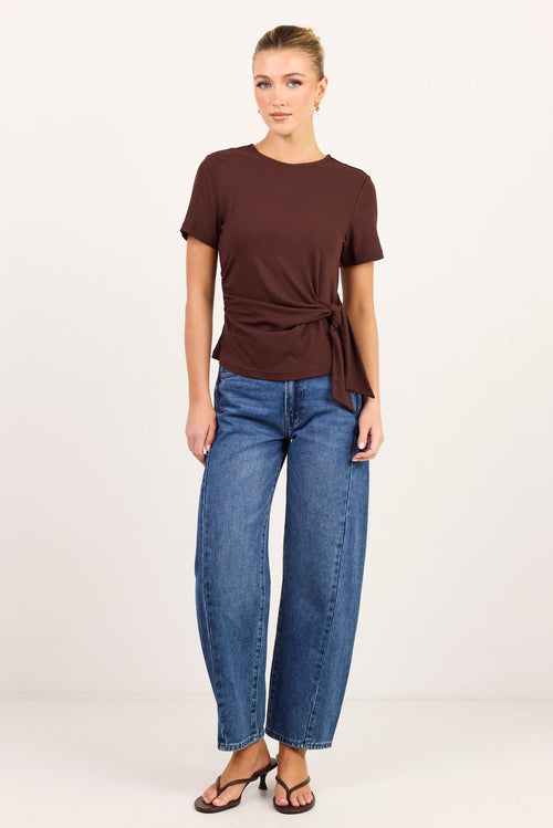 A woman modeling a dark brown short-sleeve knit top with a twisted front detail and side tie, paired with blue jeans and black sandals, against a plain white studio background.