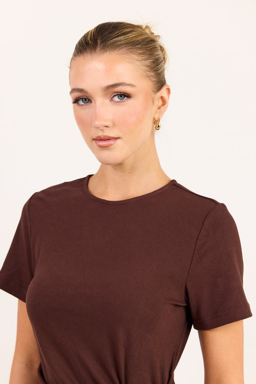 A woman with blonde hair tied back wears a simple short-sleeve crewneck t-shirt in a dark brown color, photographed against a clean off-white background.