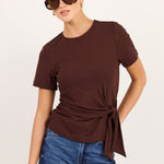 A woman modeling a dark brown short-sleeve t-shirt with a side twist detail, paired with blue jeans and sunglasses, against a plain light background.