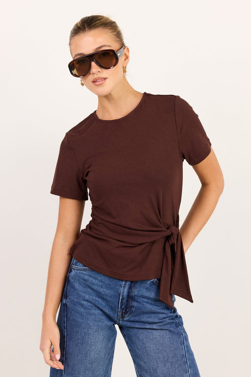 A woman modeling a dark brown short-sleeve t-shirt with a side twist detail, paired with blue jeans and sunglasses, against a plain light background.