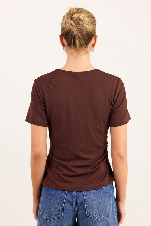 A woman viewed from the back wearing a dark brown short-sleeve t-shirt and blue jeans against a plain off-white background.