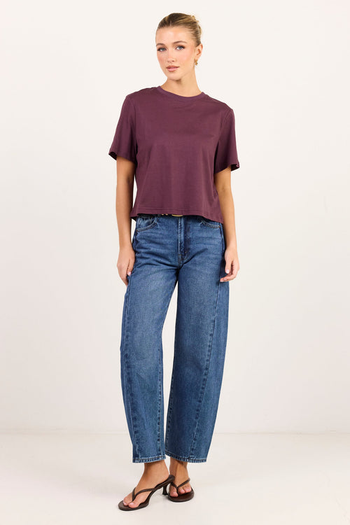 A woman modeling a short-sleeve crewneck t-shirt in deep plum purple and blue wide-leg jeans, standing against a plain white studio background.