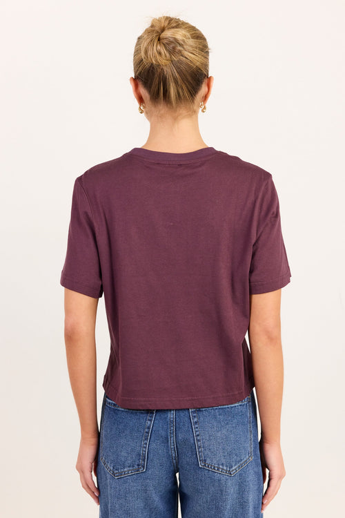 A person shown from the back wearing a short-sleeve crew-neck t-shirt in a muted plum purple color, paired with blue jeans, against a plain off-white background.