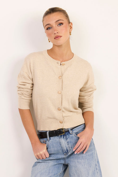 A woman modeling a beige knit cardigan with three-quarter sleeves and a button-up front, paired with light-wash blue jeans and a black belt, against a plain white studio background.