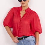 A woman modeling a red short-sleeve blouse with a ruffled collar and button front, paired with light blue jeans and black sunglasses, against a plain white background.