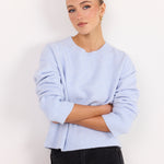A woman wearing a light periwinkle blue ribbed knit sweater with black pants, standing against a plain white studio background.