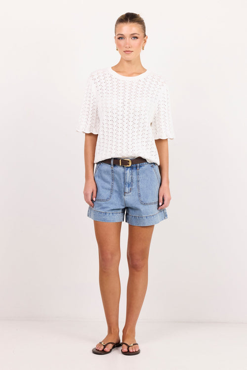 A woman modeling a white textured short-sleeve top and light blue denim shorts with a brown belt, standing against a plain white studio background.