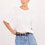 A woman modeling a white short-sleeve knit top and light blue denim shorts against a plain white studio background.