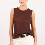 A woman modeling a sleeveless dark brown knit top paired with off-white shorts and a black belt with a gold buckle, set against a plain light gray studio background.