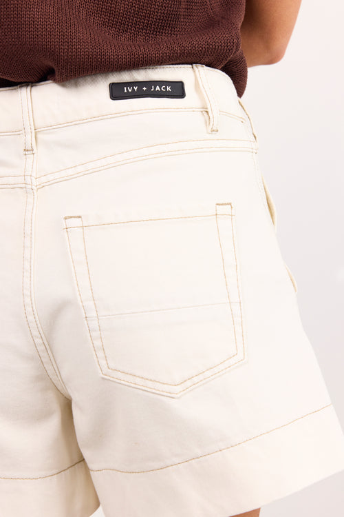 Close-up of a person wearing cream-colored denim shorts with contrast stitching and a black brand label reading 'Ivy + Jack' at the waistband, paired with a brown ribbed top against a light neutral background.