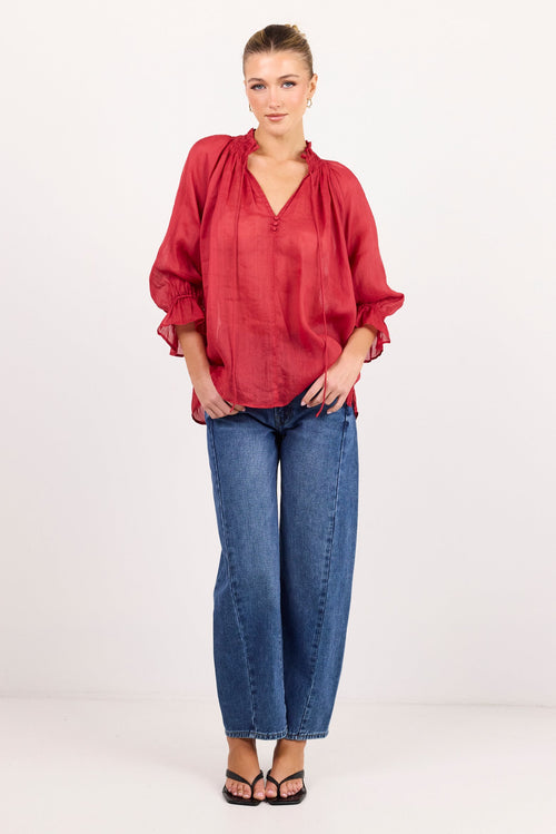 A woman modeling a loose-fitting, long-sleeved red blouse with a gathered neckline, paired with blue jeans and black sandals, standing against a plain white studio background.