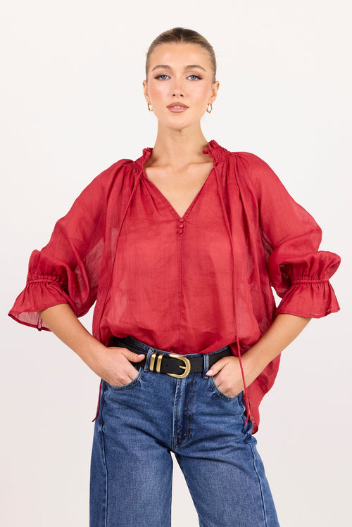 A woman modeling a red sheer long-sleeve blouse with a V-neckline and ruffled cuffs, paired with blue jeans and a black belt, against a plain light background.
