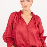 A woman modeling a red long-sleeve blouse with a gathered V-neckline and ruffled cuffs, paired with blue jeans and a black belt, against a plain white background.