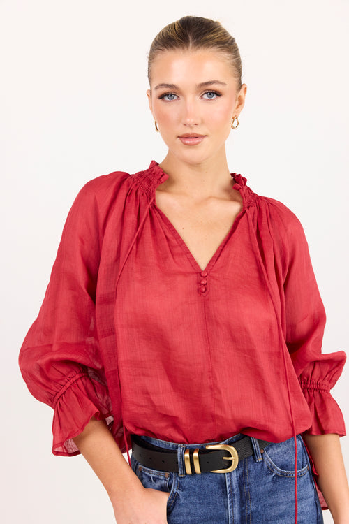 A woman modeling a red long-sleeve blouse with a gathered V-neckline and ruffled cuffs, paired with blue jeans and a black belt, against a plain white background.
