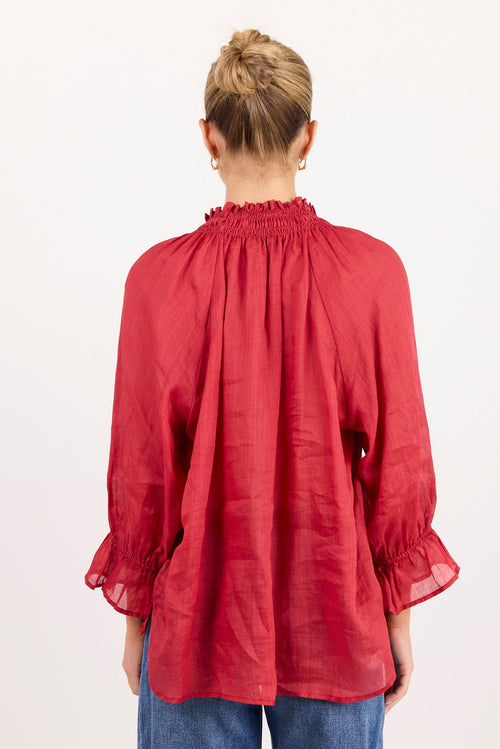 A woman viewed from the back wearing a red long-sleeve blouse with a smocked neck and ruffled cuffs, paired with blue jeans, against a plain white background.