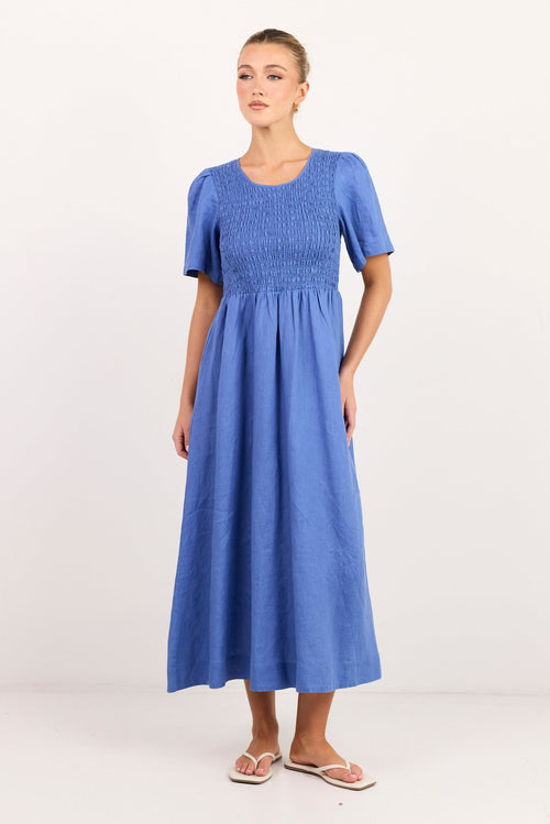 A woman modeling a blue smocked midi dress with short puff sleeves and a gathered skirt, standing against a plain white studio background.