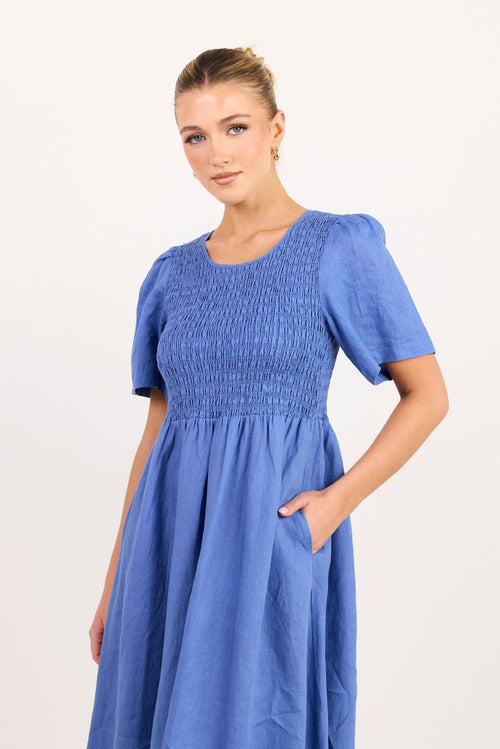 A woman modeling a blue smocked dress with short puff sleeves and a gathered skirt, standing against a plain light background.