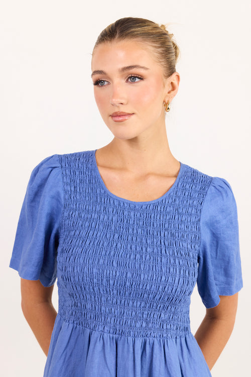 A woman wearing a blue smocked dress with short puff sleeves and a round neckline, photographed against a plain light background.