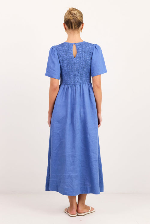 A woman viewed from the back wearing a blue midi dress with short puff sleeves, a smocked bodice, and a keyhole back detail, standing against a plain white studio background.