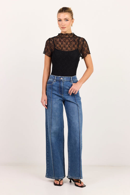 A model stands against a plain white studio background wearing a black short-sleeve top with a sheer lace overlay and high neck, paired with high-waisted, wide-leg blue jeans and black sandals.