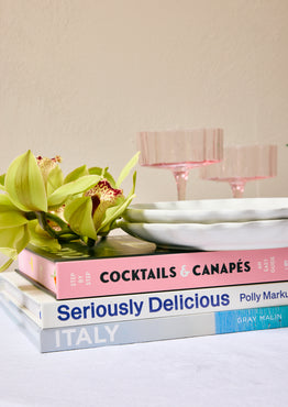 Stack of cookbooks titled 'Cocktails & Canapes', 'Seriously Delicious', and 'Italy' on a surface with a decorative plate and flowers.