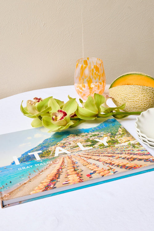 Book titled 'Italy' with a scenic cover on a table, accompanied by green orchids and a decorative hat.