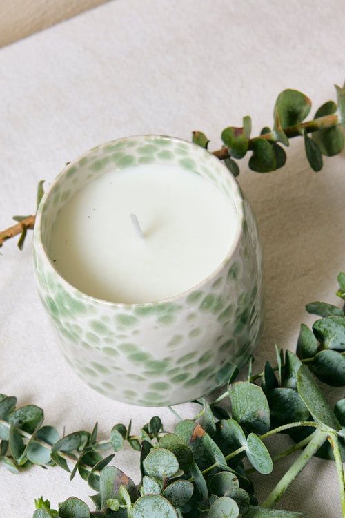 Candle in a green container surrounded by eucalyptus leaves on a light surface