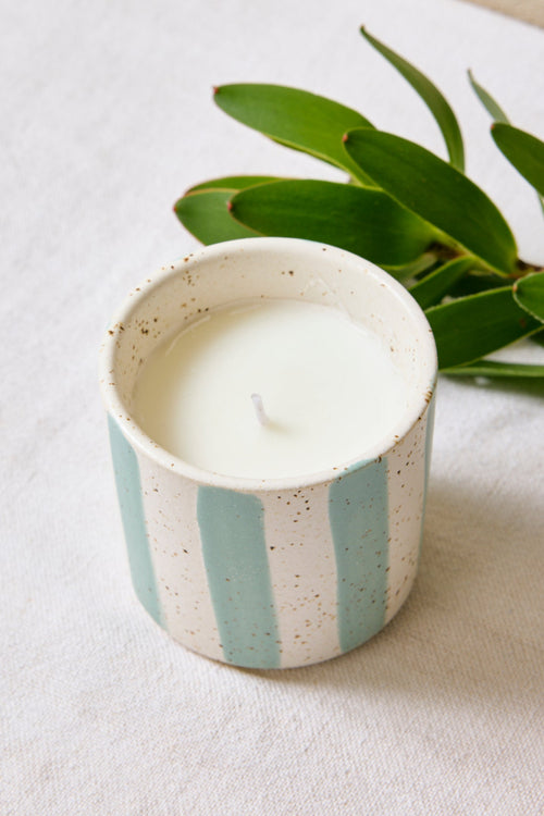 Candle in a striped ceramic holder with green leaves on a light surface