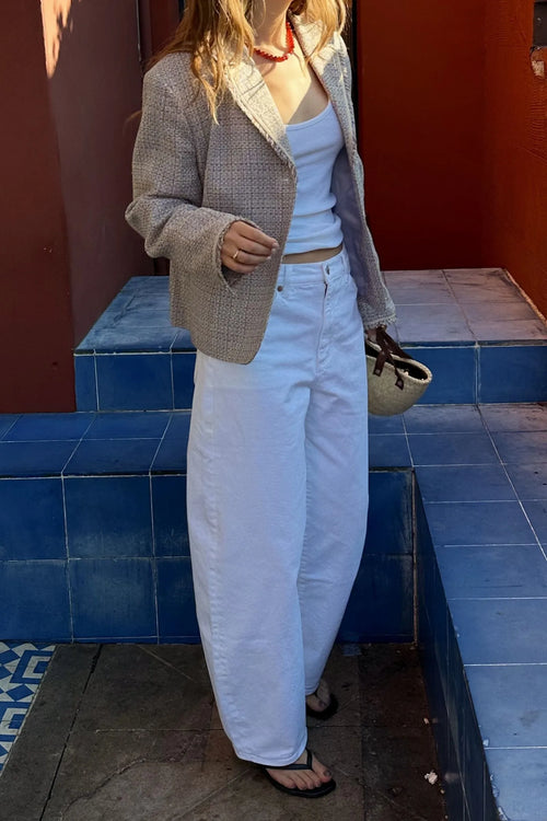 A woman wearing a beige textured tweed blazer, white tank top, and white wide-leg jeans, standing on blue tiled steps against a terracotta wall.