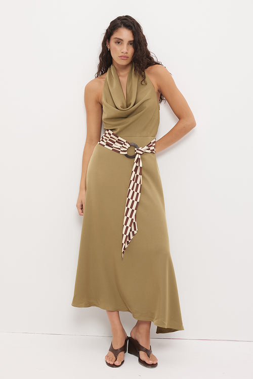 A woman modeling a sleeveless olive green maxi dress with a draped cowl neckline and a patterned brown and white tie belt, standing against a plain white studio background.