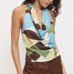 A woman wearing a sleeveless halter-neck top with a draped cowl neckline and an abstract geometric print in light blue, brown, olive green, and cream, paired with a brown fringed skirt against a plain white background.