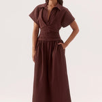 A woman modeling a dark brown midi dress with short flutter sleeves, a collared neckline, a gathered waist, and a flowing skirt, set against a plain light gray studio background.