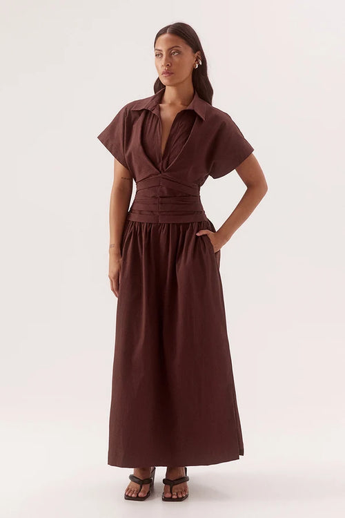 A woman modeling a dark brown midi dress with short flutter sleeves, a collared neckline, a gathered waist, and a flowing skirt, set against a plain light gray studio background.