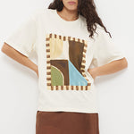 A woman modeling a cream-colored oversized t-shirt with a colorful geometric graphic print on the front, paired with a flowing brown maxi skirt and flat sandals, set against a plain white studio background.
