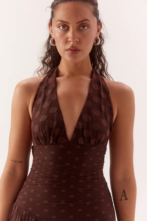 A woman wearing a dark brown halter-neck dress with a matching polka-dot pattern, photographed against a clean off-white studio background.