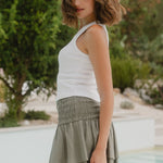 Woman wearing a white top and green skirt standing by a pool with greenery in the background