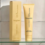 A yellow tube of Huxter hand cream in the scent "Lily & Cotton Flower" stands beside its matching rectangular box on a clear glass shelf, against a soft-tiled beige wall with natural light casting gentle shadows.