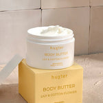 An open white jar of Huxter body butter in the scent "Lily & Cotton Flower" rests on top of its soft yellow box, revealing a smooth, whipped cream texture inside. The setup sits on a warm-toned surface against a tiled beige wall, bathed in natural light and soft shadows.