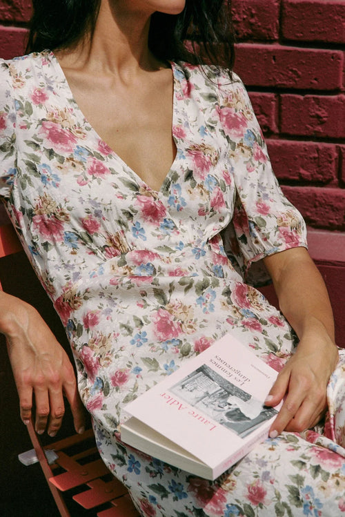 A woman wearing a white short-sleeve V-neck dress with a pink, blue, and green floral print sits on a wooden bench against a red brick wall, holding a pink book.