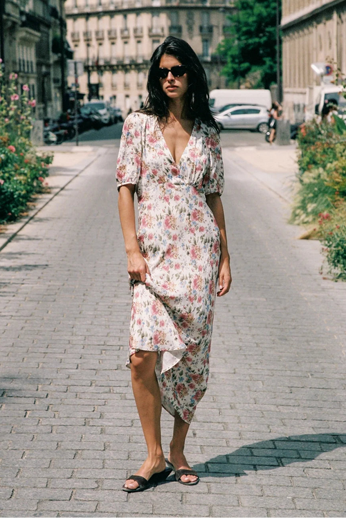A woman wearing a short-sleeve floral midi dress and black slide sandals walks down a cobblestone city street lined with greenery and buildings.