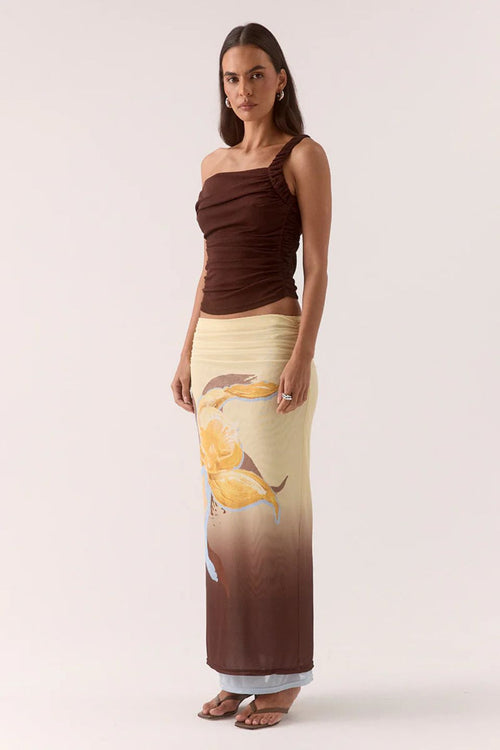 Woman wearing a brown one-shoulder top and a long, patterned skirt on a white background