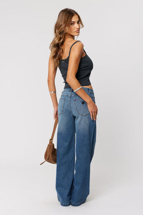 Woman wearing a black tank top and blue jeans holding a brown handbag on a white background