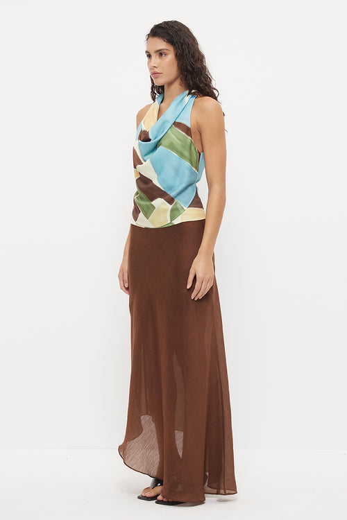 A woman modeling a sleeveless wrap-style top with a multicolor geometric print in blue, green, brown, and beige, paired with a flowing brown maxi skirt, against a plain white studio background.