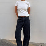 A person stands against a light gray wall wearing a fitted white short-sleeve T-shirt, loose black jeans with a studded belt, black flats, and black sunglasses.