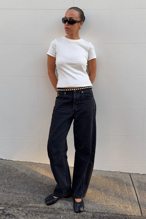 A person stands against a light gray wall wearing a fitted white short-sleeve T-shirt, loose black jeans with a studded belt, black flats, and black sunglasses.