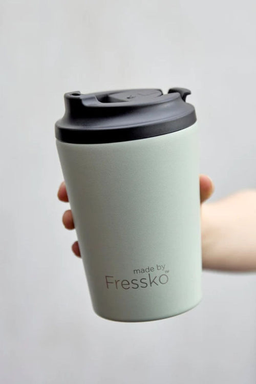 Light green coffee cup with black lid held by a hand on a light gray background, featuring 'made by Fressko' branding.