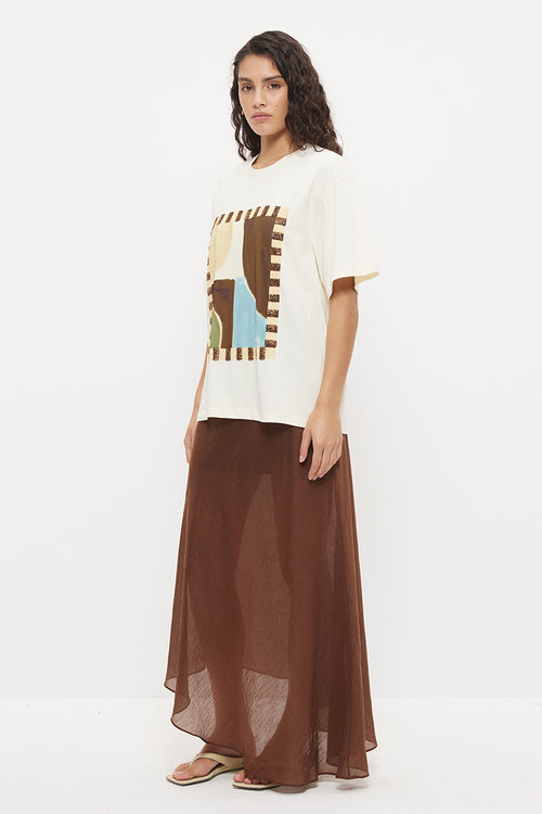 A woman modeling a cream-colored graphic t-shirt with a brown abstract print and a long brown sheer maxi skirt, standing against a plain white studio background.