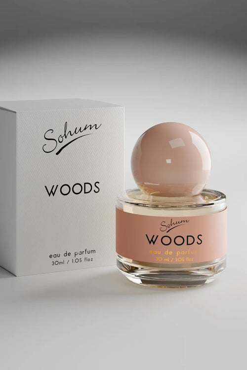A round glass bottle of Sohum Woods eau de parfum with a glossy nude-toned spherical cap and a warm terracotta-pink label, featuring black and mustard-yellow text for an earthy, sophisticated aesthetic.