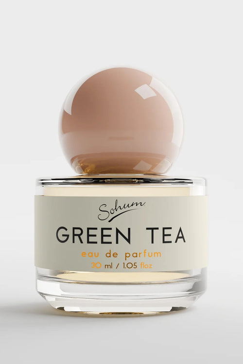 A round glass bottle of Sohum Green Tea eau de parfum with a soft blush-toned spherical lid, placed beside its minimal white textured box. The label is cream with black and amber lettering.